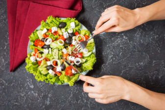 Traditional greek salad with fresh vegetables, feta cheese and olives on stone background. Female hands hold knife and firk