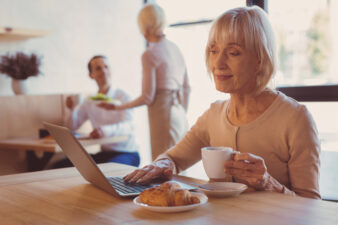 Older woman on laptop eating croissant drinking coffee