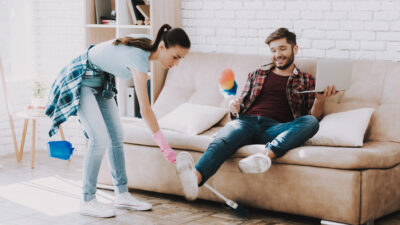 Woman cleaning under the feet of man sitting on couch