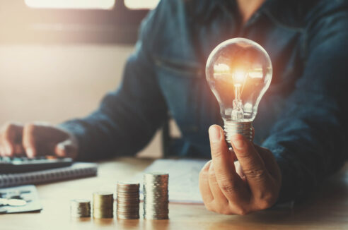 man holding up a lightbulb while looking at stack of coins