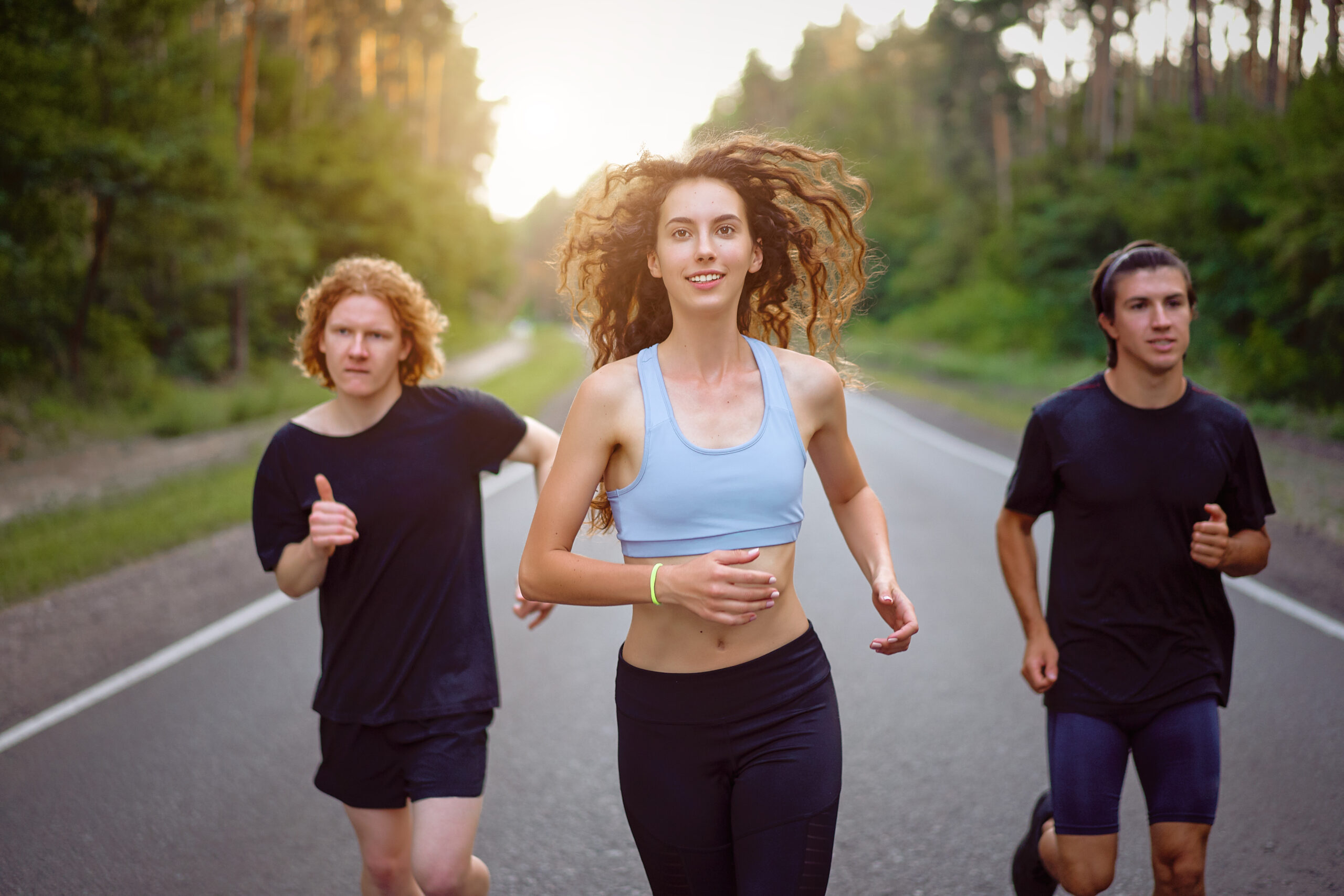 A group of three people athletes one girl and two men run on an asphalt road in a pine forest