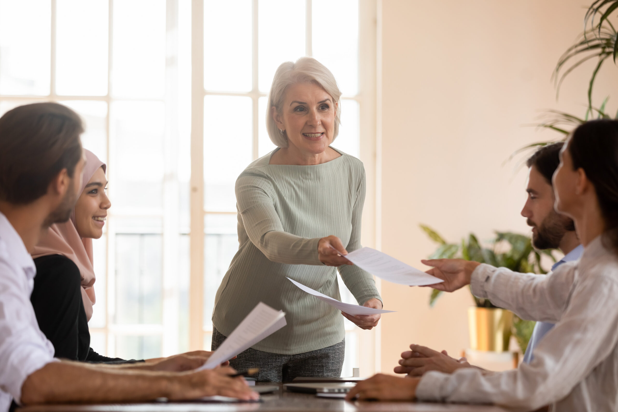 Older female leader give papers to diverse team at briefing