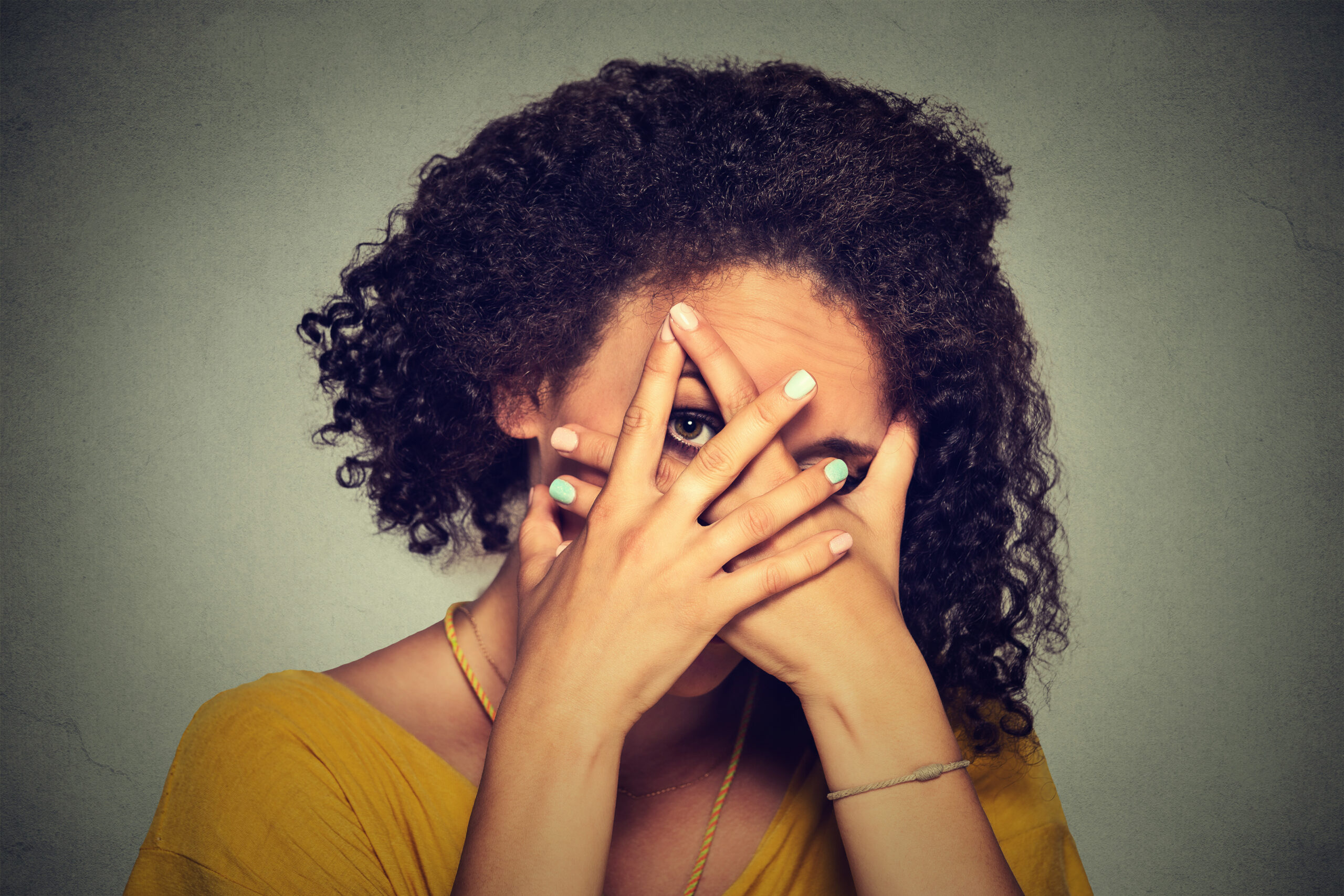 Shy woman hiding face timid. Woman peeking though hands. Gray background.