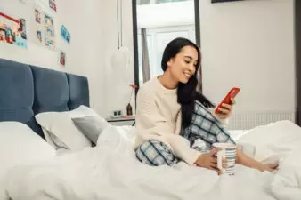 Smiling woman sitting in bed with phone and coffee