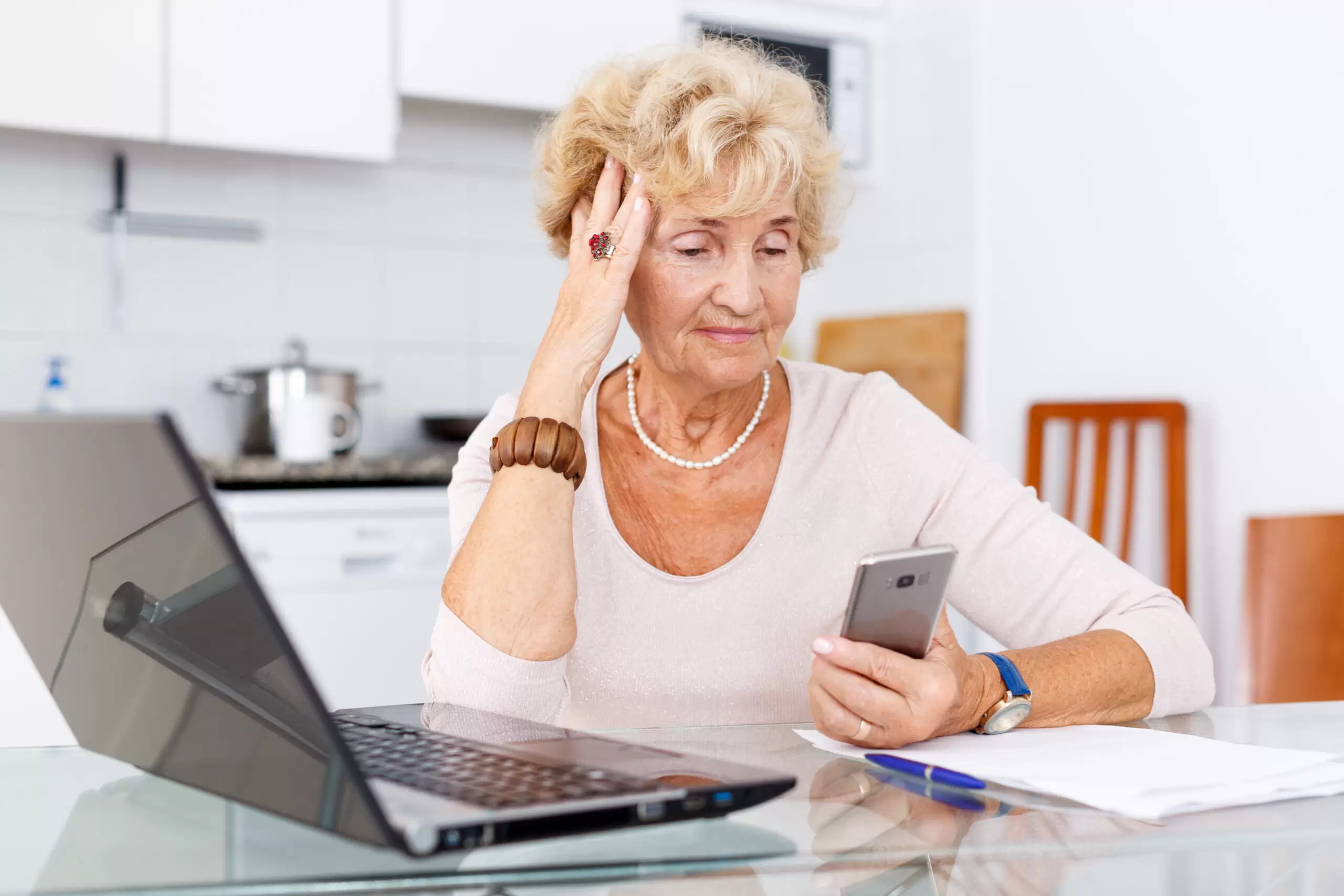 Confused elderly woman using laptop and smartphone at kitchen table