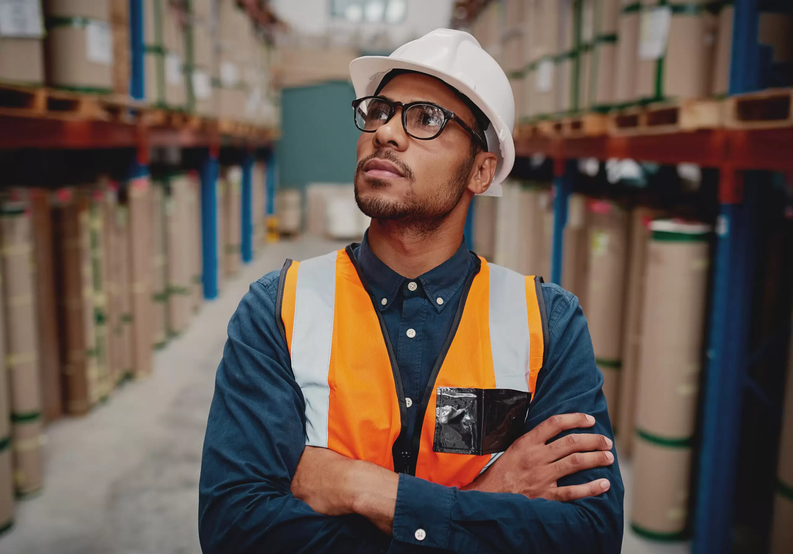 Portrait of serious supervisor in warehouse standing with folded hands looking high at shelves with material while wearing orange vest and white hardhat