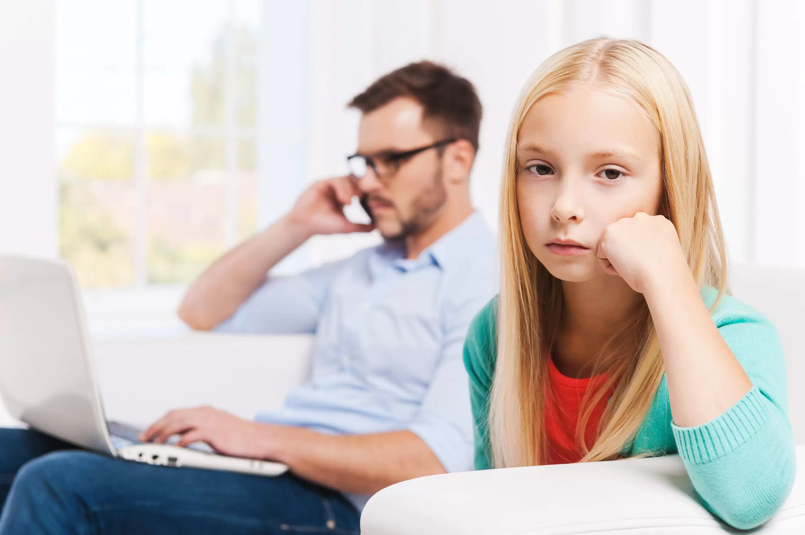 Feeling lonely at those moments. Busy young man working on laptop and talking on the mobile phone with his daughter sitting on foreground and looking upset