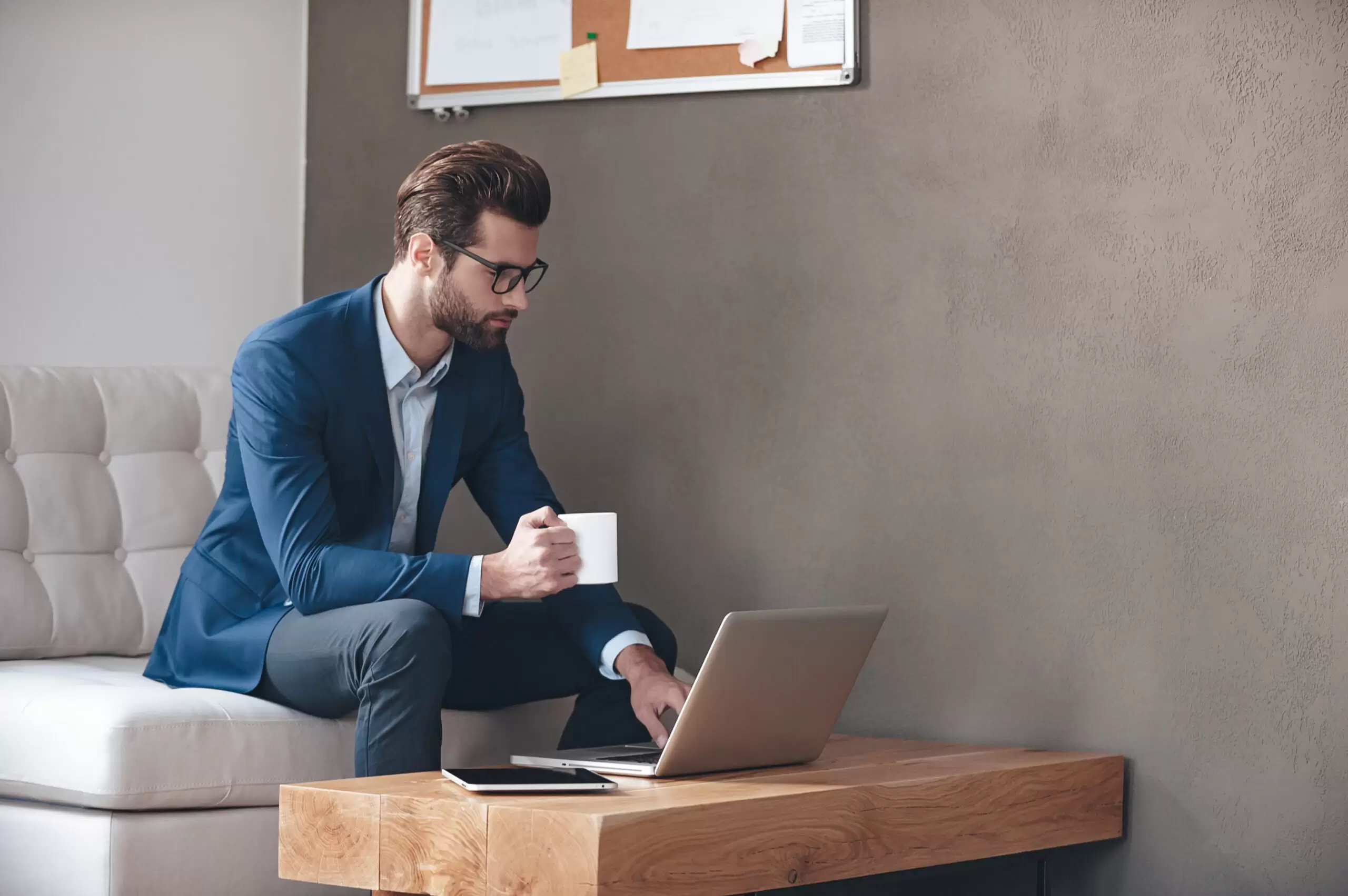 Starting new working day. Handsome young man wearing glasses holding coffee cup and working with laptop while sitting on the couch in office