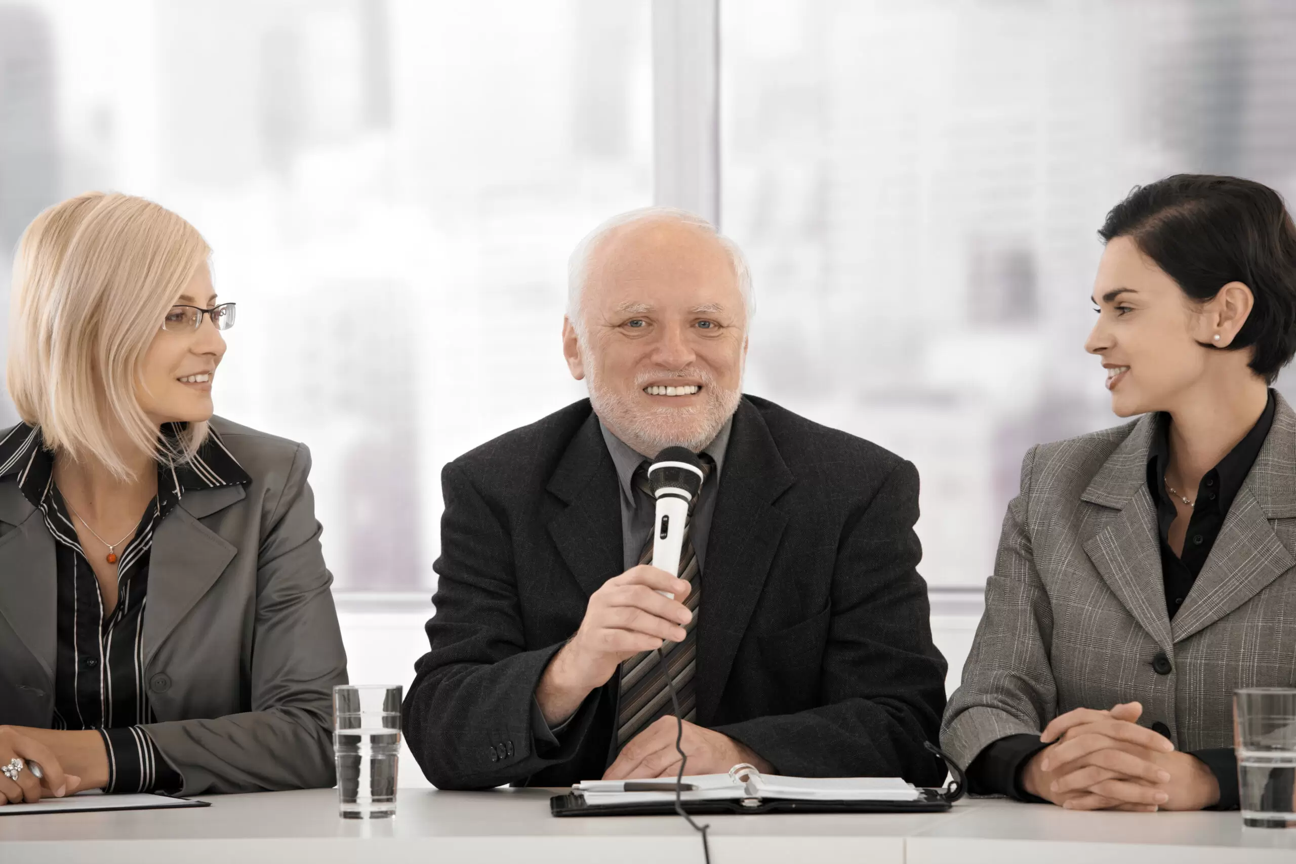 Senior businessman speaking into microphone, female colleagues watching, smiling.